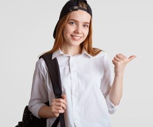 teen girl wearing white button down shirt with backpack over shoulder and backwards baseball cap